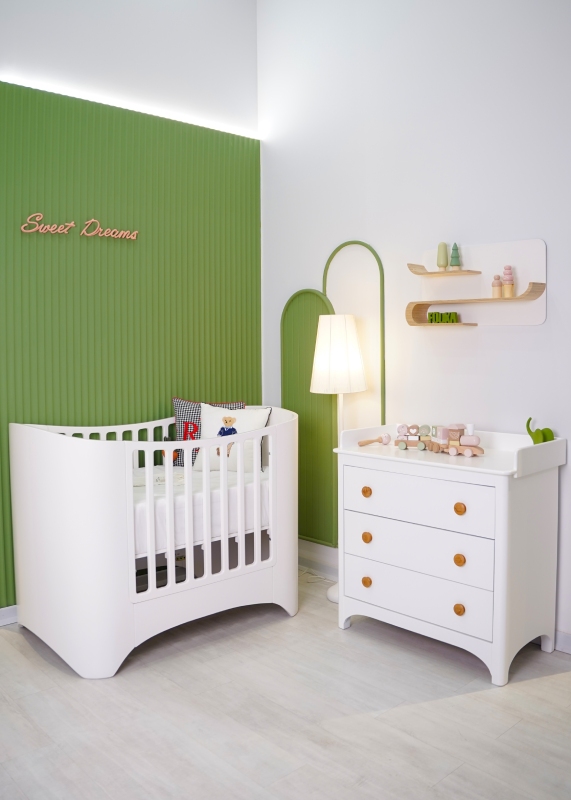 نمای داخلی اتاق نوزاد با تخت و دراور سفید سوییت و نورپردازی ملایم کنار دیوار سبز شیاردار